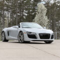 White Audi R8 Spyder convertible parked on a concrete driveway with trees in the background, basketball hoop to the left menioned.