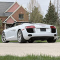 White Audi R8 convertible parked on a driveway with a brick house and trees in the background, daytime scene.